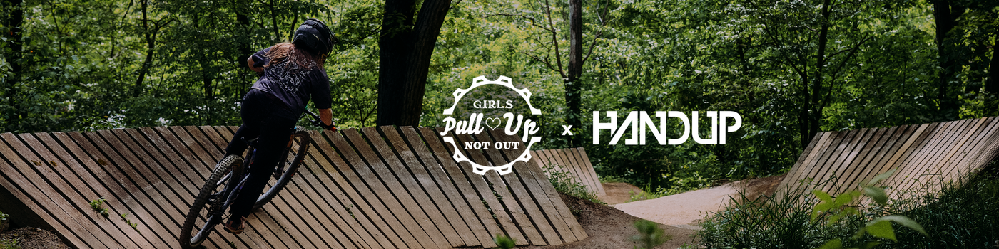 A mountain biker rides a bike over wooden ramps in a forest setting, with a central 'Girls Pull Up Not Out' logo and a 'HANDUP' brand mark overlaid on the scene.