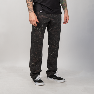 A person wearing black jeans with a subtle dark gray marble or distressed pattern, styled with a black t-shirt and black low-top sneakers with white laces, standing against a light gray studio background.